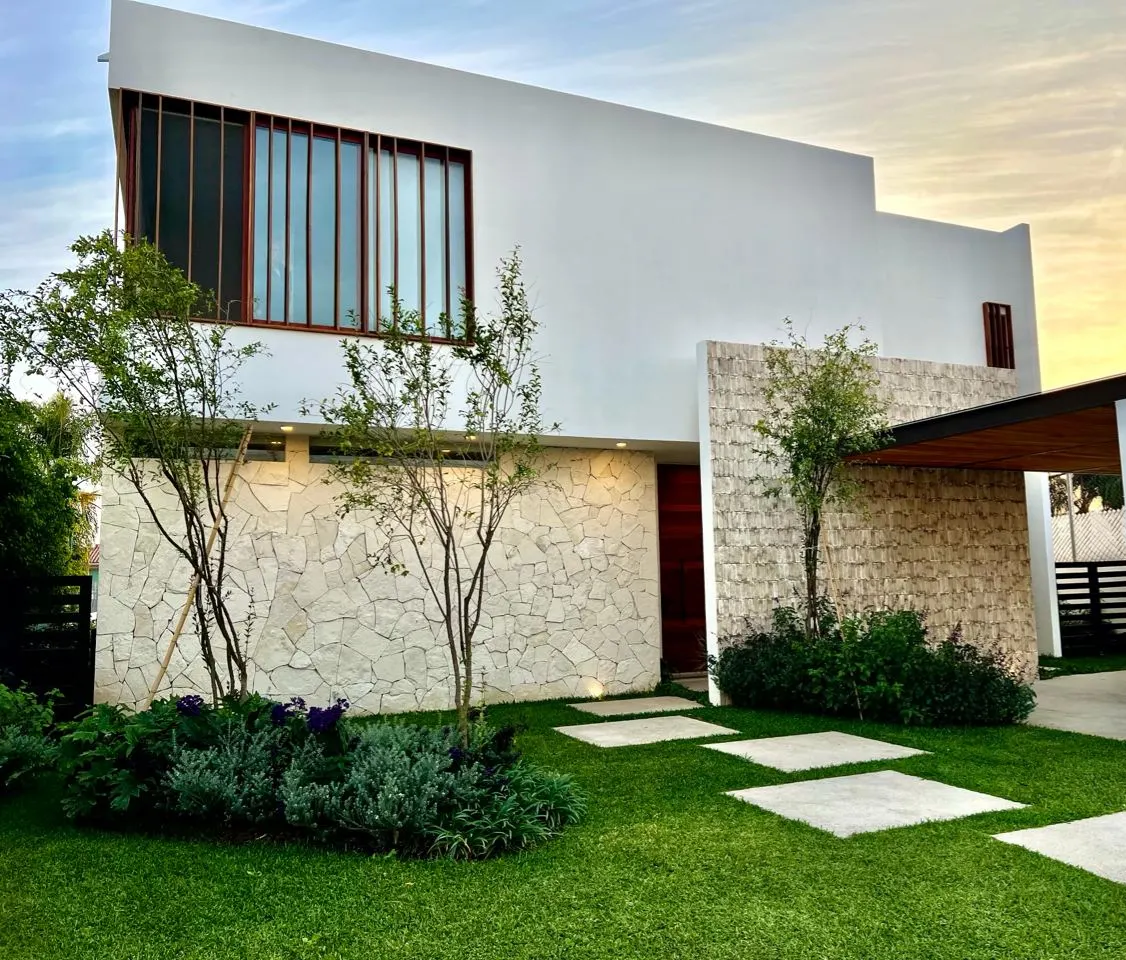 Fachada de la casa al atardecer, con muro de piedra, celosía de madera y jardín delantero.